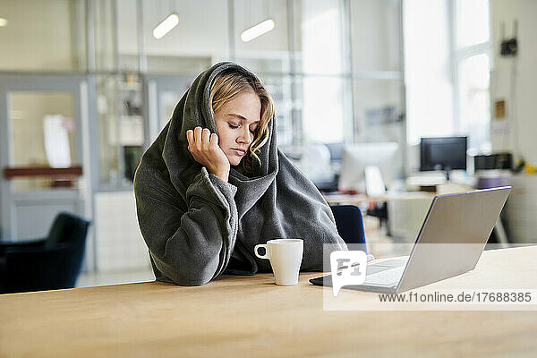 Exhausted young woman in cozy loungewear sitting at desk in office with laptop