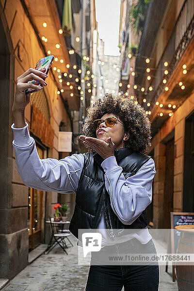 Young woman blowing kiss taking selfie through mobile phone in alley