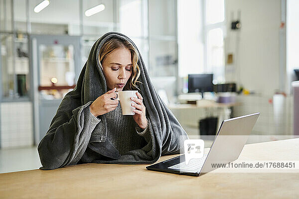 Young woman in cozy loungewear sitting at desk in office drinking from cup