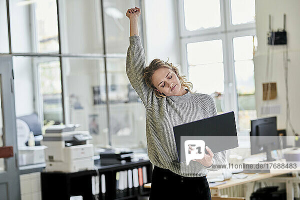 Tired young woman standing in office holding laptop