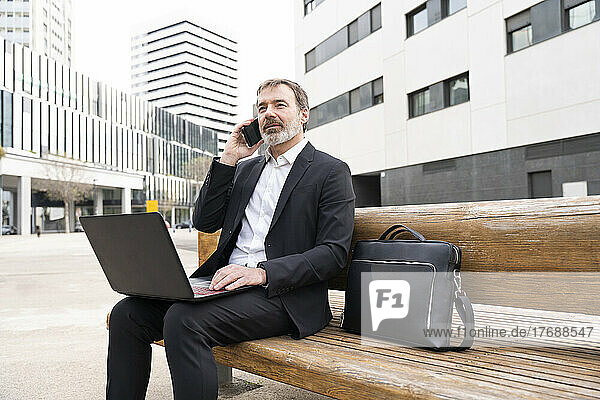 Mature businessman talking on mobile phone sitting with laptop on bench
