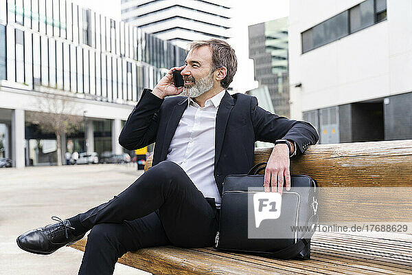 Smiling businessman talking on smart phone sitting with laptop bag on bench
