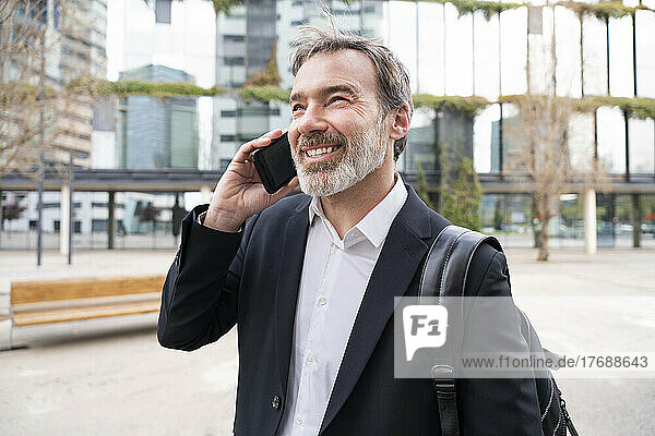 Glücklicher Geschäftsmann mit Rucksack  der auf dem Fußweg mit dem Smartphone spricht