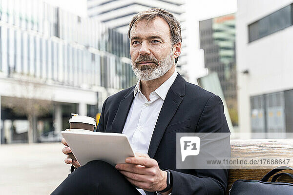 Mature businessman with tablet PC and disposable coffee cup sitting on bench