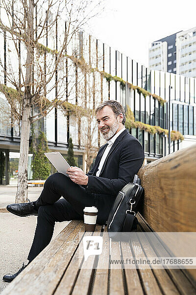 Smiling businessman with tablet PC sitting by laptop bag on bench