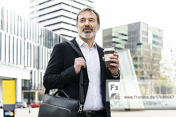 Smiling mature businessman with laptop bag holding disposable coffee cup