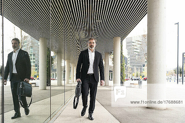 Mature businessman carrying laptop walking by glass wall