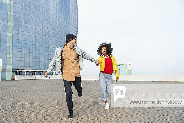 Happy couple holding hands running on footpath in front of building