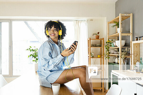 Lächelnde junge Frau mit Kopfhörern sitzt zu Hause auf der Kücheninsel