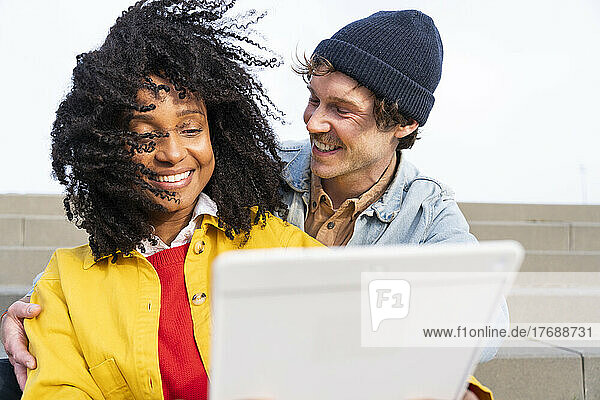 Happy man looking at girlfriend with tousled hair using tablet PC