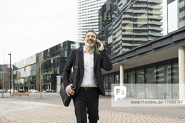 Smiling mature businessman with laptop bag talking on smart phone walking outside office building