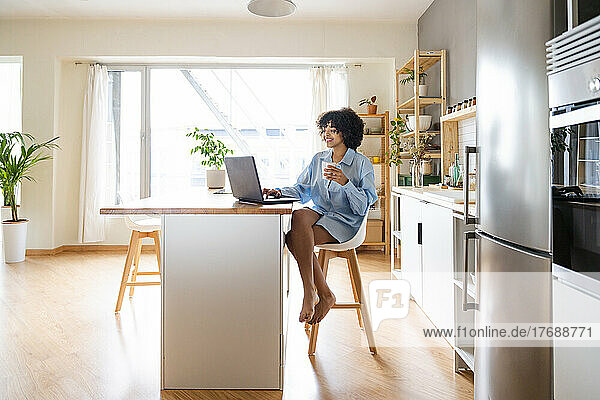 Happy woman using laptop sitting on chair at kitchen island