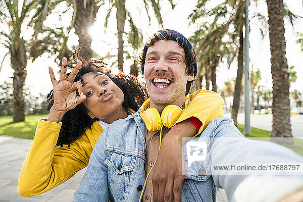 Fröhlicher Mann mit Kopfhörern neben Frau  die im Park ein „OK“-Schild zeigt