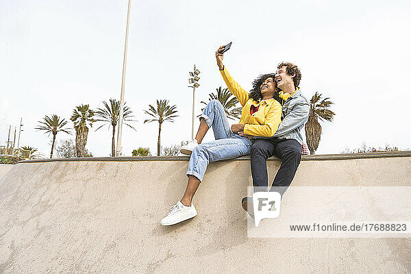Cheerful couple taking selfie through mobile phone sitting on wall