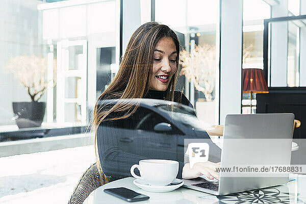 Smiling businesswoman using laptop seen through glass window in cafe