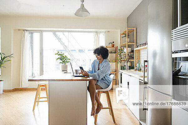 Smiling woman with smart phone using laptop sitting on chair at kitchen island