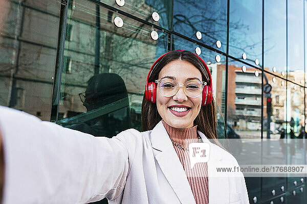 Happy woman wearing wireless headphones standing in front of glass wall