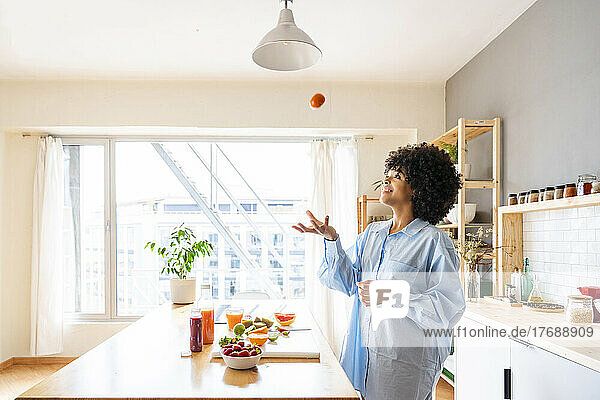 Beautiful woman catching tomato standing by kitchen island at home