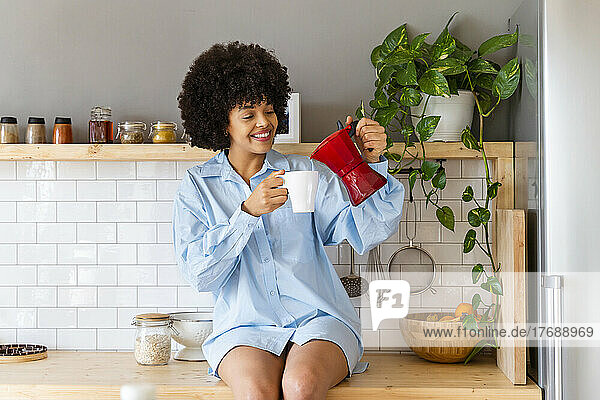 Glückliche Frau  die zu Hause auf der Küchentheke sitzt und Kaffee in eine Tasse gießt