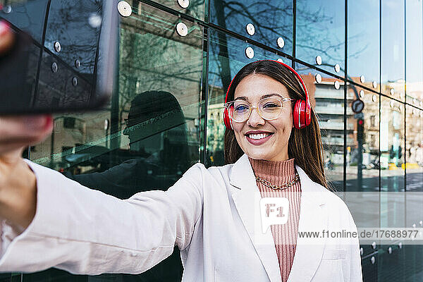Smiling woman wearing wireless headphones taking selfie through smart phone