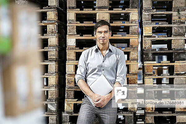 Confident businessman with laptop in front of stack of pallets at warehouse