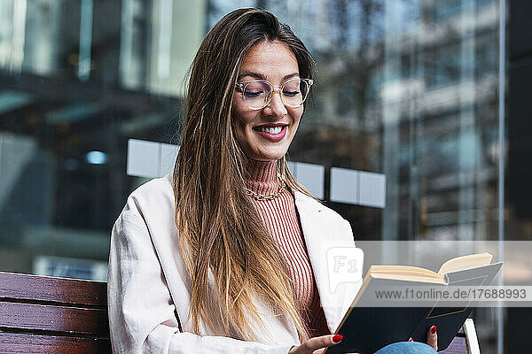 Smiling woman wearing eyeglasses reading book sitting in front of glass wall