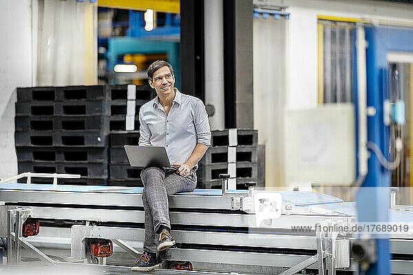 Contemplative businessman with laptop sitting on conveyor belt in factory