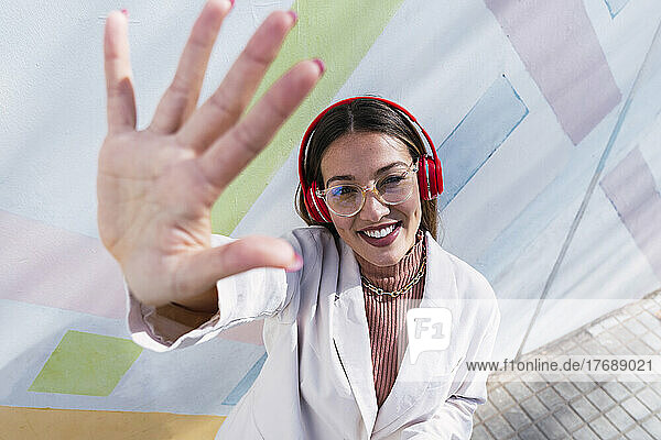 Smiling woman wearing headphones gesturing hand in front of wall on sunny day