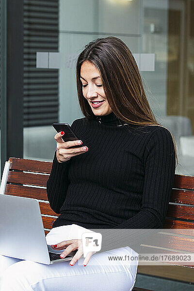 Smiling businesswoman using smart phone sitting with laptop on bench