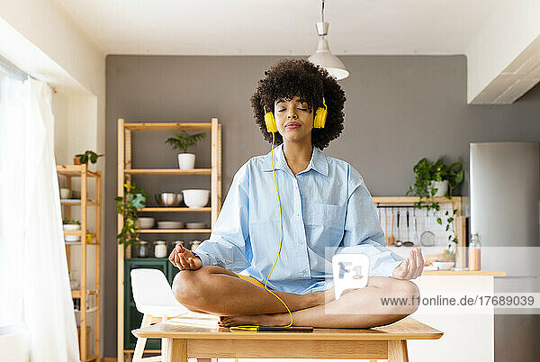 Beautiful woman wearing headphones meditating on dining table at home