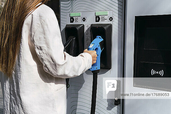 Young woman holding electric plug at charging station on sunny day