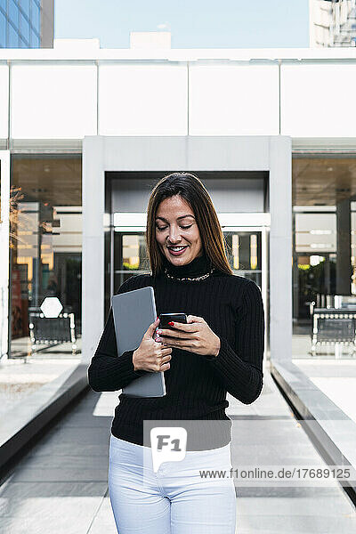 Smiling young businesswoman with laptop using smart phone in front of cafe