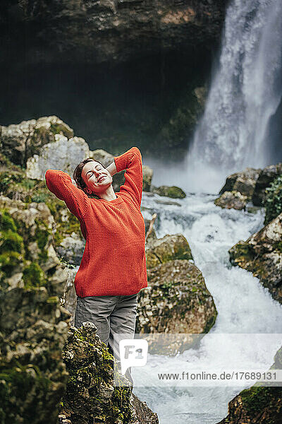 Glückliche junge Frau mit den Händen hinter dem Kopf  die vor dem Wasserfall steht