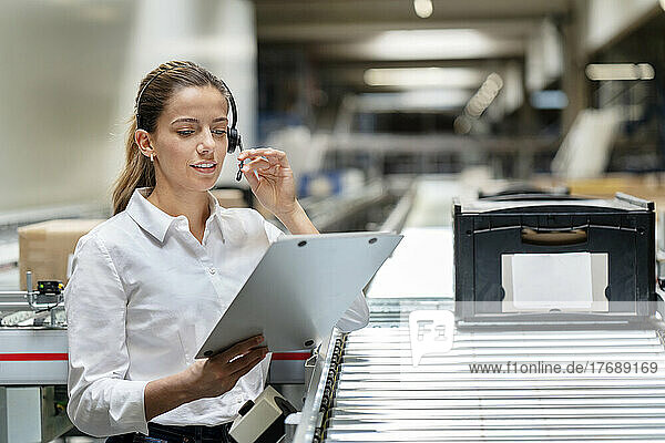 Young businesswoman reading on clipboard with headset at factory
