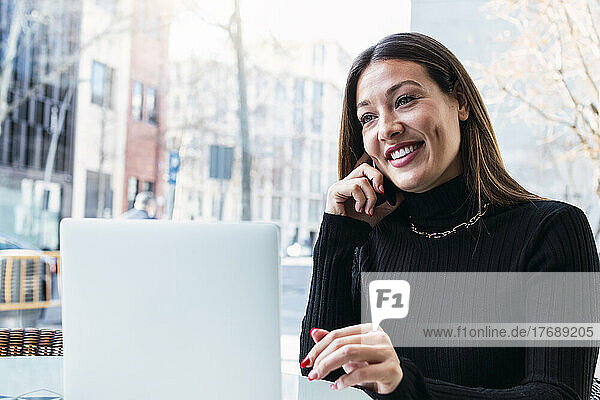 Lächelnde junge Geschäftsfrau mit Laptop  die im Café mit dem Handy telefoniert
