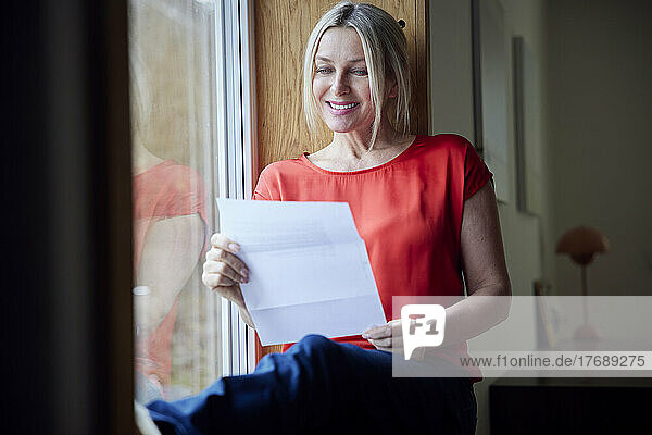 Smiling woman reading letter sitting by window at home