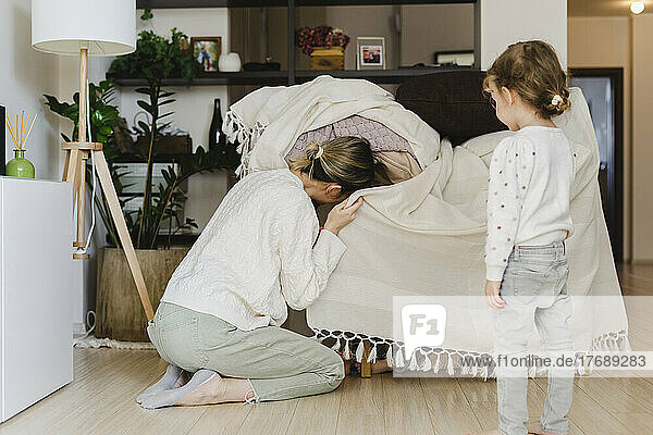Playful mother peeking in blanket by daughter standing at home
