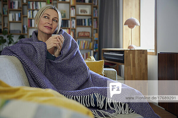 Mature woman wrapped in blanket sitting on sofa day dreaming at home