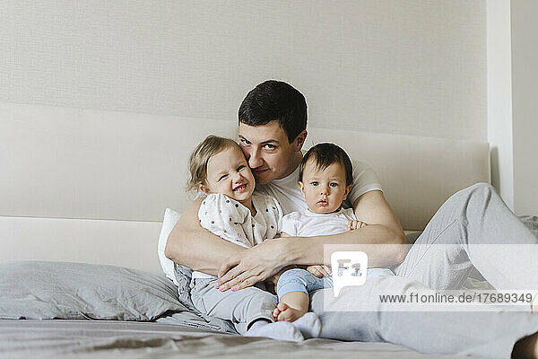 Smiling man embracing son and daughter sitting on bed at home