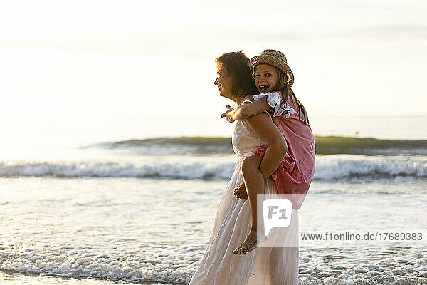 Smiling mother giving piggyback ride to cheerful daughter at beach
