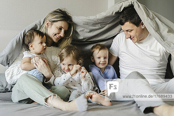 Happy parents with children sitting under blanket in bedroom