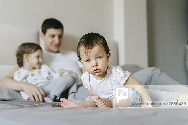 Cute baby boy with father and sister in background on bed at home