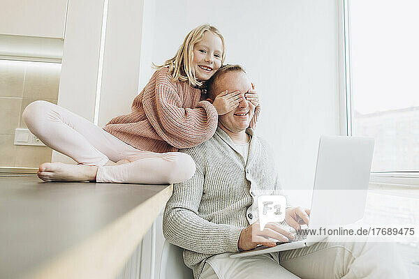 Glückliche Tochter bedeckt die Augen ihres Vaters  der zu Hause mit Laptop am Fenster sitzt