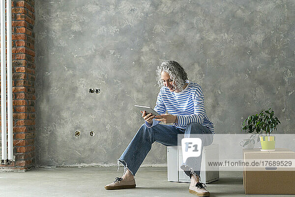 Smiling woman with gray hair using tablet PC sitting on box in front of wall