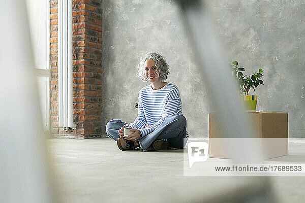 Happy woman sitting on ground with cup in front of wall