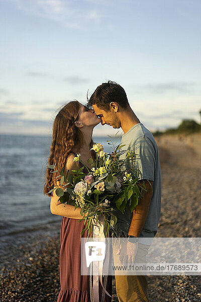 Frau mit Blumenstrauß küsst Mann am Strand auf die Stirn