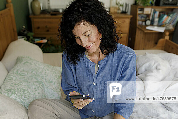 Smiling woman using mobile phone sitting on bed