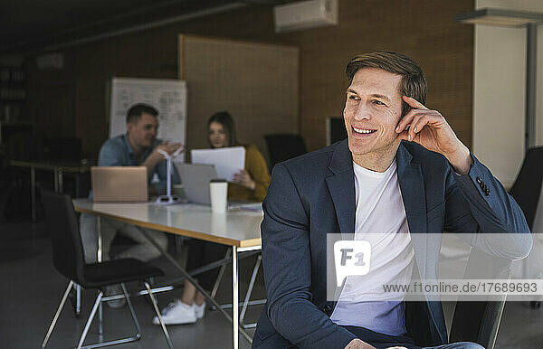 Confident businessman sitting on chair in office with colleagues in background