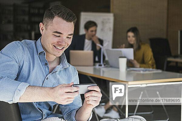 Smiling businessman using smartphone in office with colleagues in background