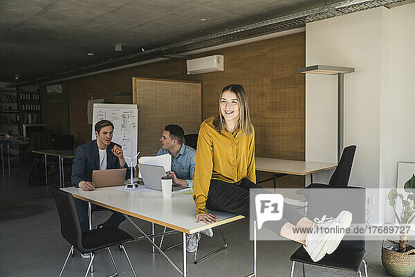 Happy businesswoman sitting on table in office with colleagues in background